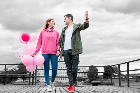 Walking and talking. Cheerful relaxed young girl holding a bunch of pink balloons while walking on the pier with her interesting handsome boyfriendの写真素材