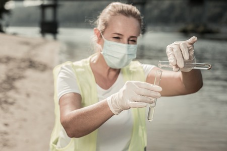 Test tubes. Sanitary inspector wearing gloves and mask holding test tubes with river water while...
