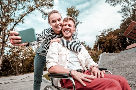 Funny selfie. Positive happy young man sitting in the wheelchair and laughing while loving woman...