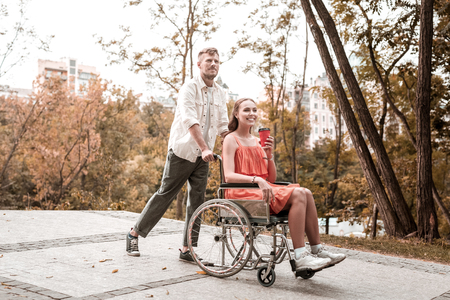 Walk in park. Cheerful wheelchair girl drinking hot coffee and smiling while spending time in the park with positive boyfriendの写真素材