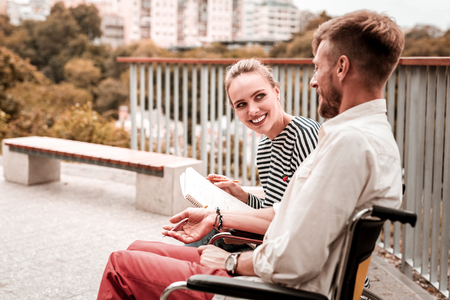 Curious smile. Cheerful young woman holding big notebook and looking friendly while smiling to the disabled friendの写真素材