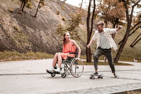 Fast penny board. Cheerful curious wheelchair user smiling and looking at her boyfriend riding his penny boardの写真素材