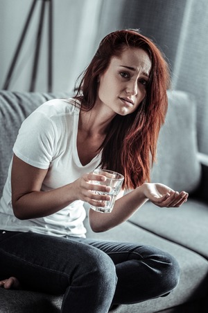 Healthcare activity. Upset longhaired young woman holding glass in right hand and wrinkling forehead while looking at cameraの写真素材