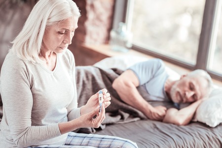 Health problems. Portrait of elderly woman looking at thermometer with sadness while her husband...