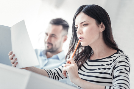 Creating plan. Pretty brunette holding pencil near mouth and looking forward while trying to concentrateの写真素材