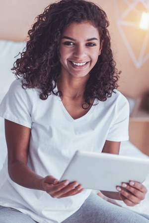 Beautiful smile. Dark-eyed pretty curly girl feeling wonderful while having beautiful broad smile spending free time at homeの写真素材