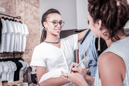 Smiling designer. Smiling dark-haired prosperous fashion designer wearing white dress feeling joyful working with partnerの写真素材