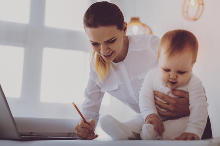 Data corrections. Financial director on maternity leave feeling occupied while correcting her business data hugging her little daughterの写真素材