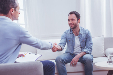 Gratitude. Pleased positive bearded man shaking hands with professional psychiatrist while being extremely thankful to him for a high quality assistanceの写真素材