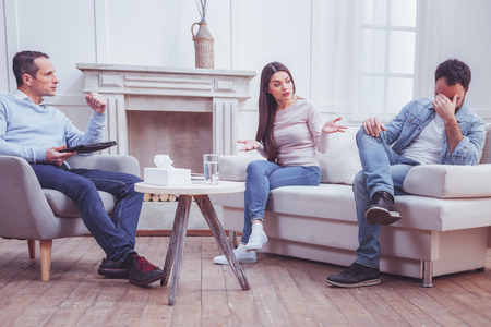 Outraged good looking young woman throwing up her hands while discovering her husbands infidelity during psychiatric attendanceの写真素材