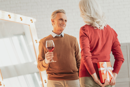 Happy elderly woman hiding a gift behind her back and preparing for congratulating her lovely husband while her husband standing in front of her with the glass of wine and smilingの写真素材