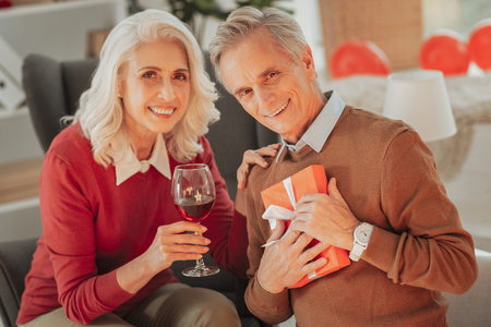Portrait of pleasant joyful senior woman putting hand on her husbands shoulder while her husband keeping the present close to his chest and expressing satisfactionの写真素材