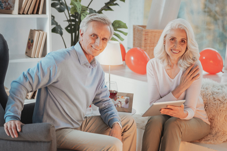 Married senior couple sitting in comfortable chairs and looking at camera with a smile while holding...