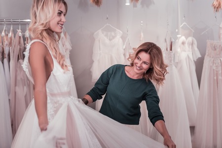 Delighted nice woman standing near her daughter while helping her with a wedding dressの写真素材