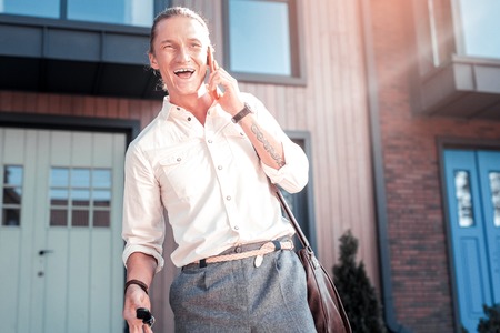 Stylish handsome man with tattoo on hand feeling very cheerful laughing while speaking by phone