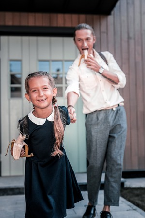 Cheerful dark-haired schoolgirl with nice hairstyle leaving house with father eating bananasの写真素材