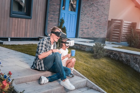 Modern father and daughter wearing virtual reality glasses and eating popcorn sitting on the porchの写真素材