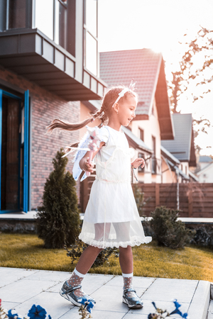Little beautiful girl wearing white dress and holding magic wand walking outside houseの写真素材