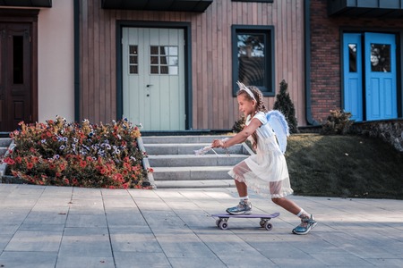 Dark-haired little beautiful girl wearing white dress and fairy wings skateboarding near houseの写真素材