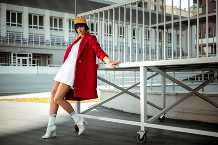 Remarkable fit model leaning on metal platform wearing fluffy red overcoat