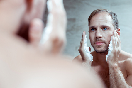 Handsome blue-eyed man using shaving foam while shaving face standing in front of mirrorの写真素材
