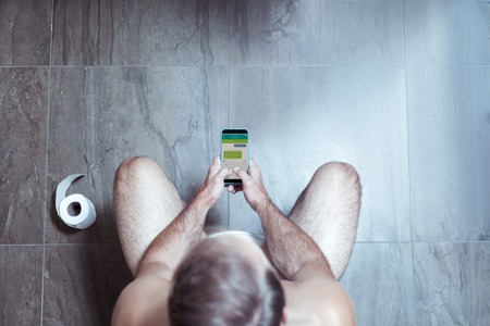 Blonde-haired man holding his smartphone and texting friends while sitting in the toiletの写真素材