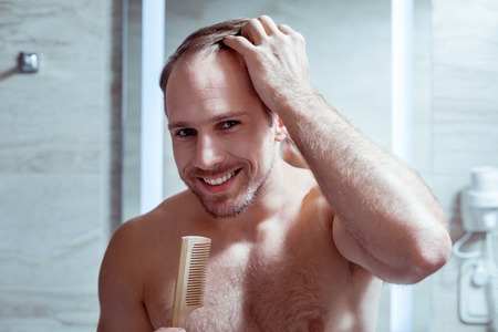 Beaming bearded blue-eyed man smiling while combing his hair standing in the bathroom