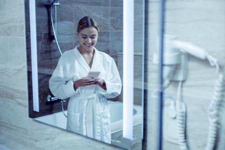 Appealing good-looking woman wearing white bathrobe reading message on her smartphoneの写真素材