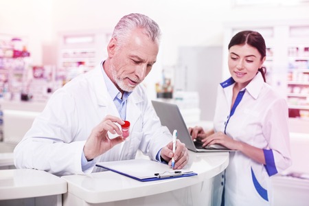 Grey-haired doctor inspecting and listing container with a medicine while curious woman working with a computerの写真素材