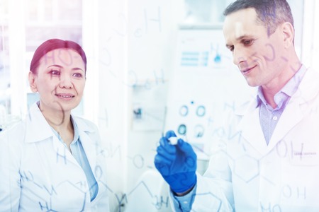 Positive cheerful man standing together with his colleague while writing a chemical formula on the whiteboardの写真素材