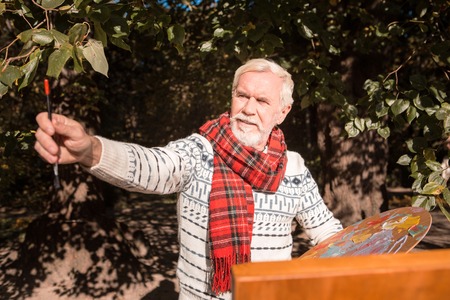 Professional artist. Joyful nice man holding his painting brush while being involved in the process of paintingの写真素材