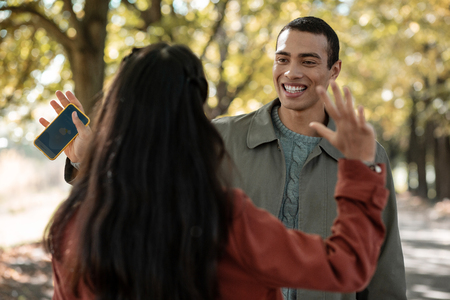 Positive delighted Asian man holding telephone in right hand while waving to his girlfriendの写真素材