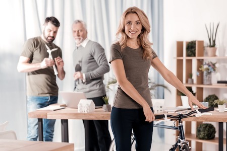Great shape. Cheerful blond woman standing with her bike while her colleagues working in the...