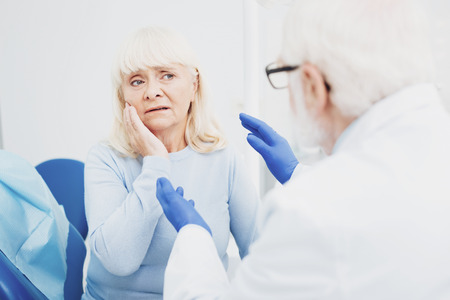 Health issue. Unhappy female patient touching her face while listening to doctorの写真素材