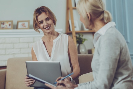 Assistance. Delighted elderly woman sitting on the couch and writing notes in notebook while having pleasant conversation with cheerful social workerの写真素材