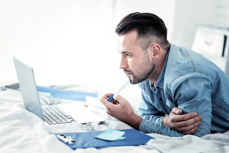 Handsome bearded man lying on elbows and touching chin with pen while working at his projectの写真素材