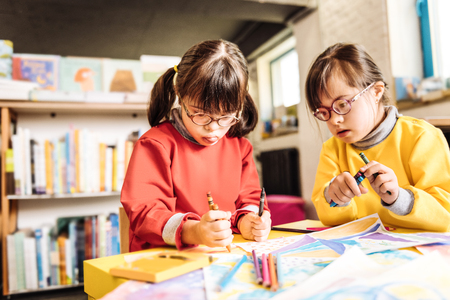 Holding crayons. Funny cute dark-haired sisters having Down syndrome holding crayons while drawingの写真素材