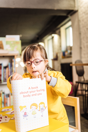 Kids book. Lovely preschool girl with genetic disorder looking at kids book sitting at the tableの写真素材