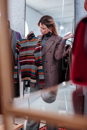 To fitting room. Appealing blonde-haired woman taking colorful striped polo neck to the fitting room