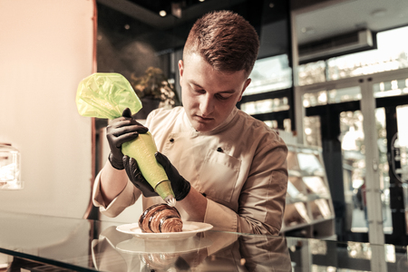 Dessert decoration. Serious professional man using a pastry bag while decorating a croissantの写真素材