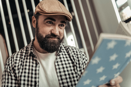 Interesting story. Joyful nice bearded man reading a book while sitting in the cafeteriaの写真素材