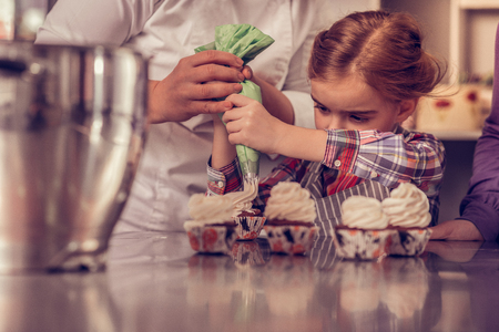 Delicious cupcakes. Pleasant creative girl holding a piping bag while decorating muffins with whipped creamの写真素材