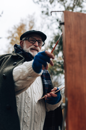 So happy. Kind grey-haired male person keeping smile on his face while drawing nature landscapeの写真素材