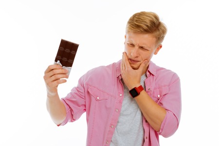 Unpleasant ache. Nice young man feeling strong tooth pain while eating a bar of chocolateの写真素材