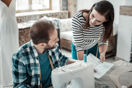 Talking to wife. Bearded husband wearing squared shirt sitting at the sewing machine talking to wifeの写真素材