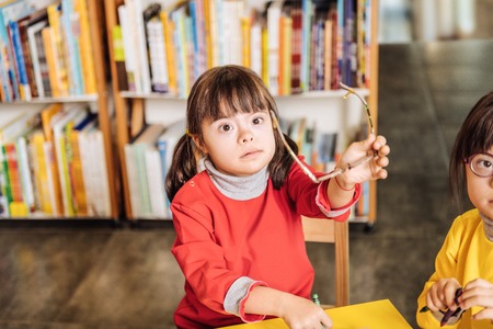Glasses off. Cute dark-eyed girl with Down syndrome taking her glasses off sitting at the table