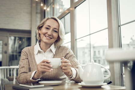 Woman with tea. Stylish mature blonde-haired woman with facial wrinkles drinking tea in cafeteriaの写真素材