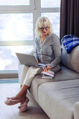 Book lover. Nice serious woman looking at the book while sitting with her laptopの写真素材