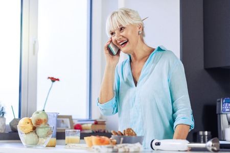 Mobile communication. Joyful happy woman talking on the phone while cooking in the kitchenの写真素材
