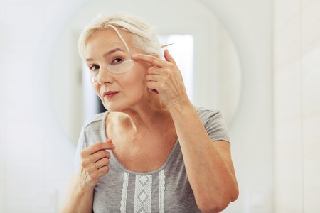 Perfect look. Nice aged woman putting eye patches on her face while standing in front of the windowの写真素材